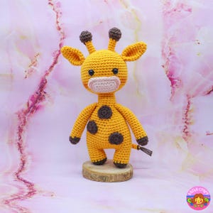 May include: A crocheted giraffe toy with brown spots, a pink nose, and brown eyes. The giraffe is standing on a wooden base.