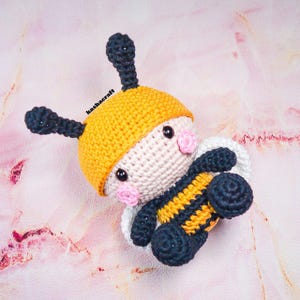 May include: A handmade crochet bee toy with a yellow head, black antennae, and a body striped with yellow and navy blue. The bee has pink cheeks and black eyes. The text "kashacraft" is visible on the top left.