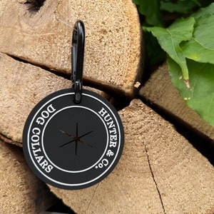 May include: Black round dog collar tag with white text that reads "DOG COLLARS" and "HUNTER & CO.". The tag has a small hole in the center and a black metal clip attached to the top.