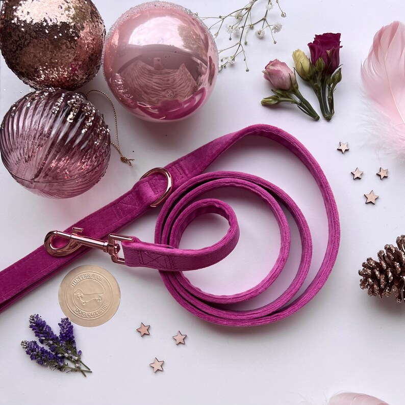 May include: A pink velvet dog leash with a gold clasp and a rose gold charm with the text "Hunter & Co. Dog Collars". The leash is coiled on a white surface with pink and gold ornaments and stars.