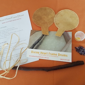 May include: A kit for making a sacred rattle, including two leather circles, a wooden stick, a roll of leather cord, a small container of purple crystals, and instructions on how to make your own sacred rattle. The instructions include the text "Divine Heart Frame Drums Make Your Own Sacred Rattle".