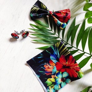 May include: A set of men's accessories featuring a black bow tie with a tropical floral print, a pair of cufflinks with a red and black design, and a matching pocket square with a vibrant tropical floral print.