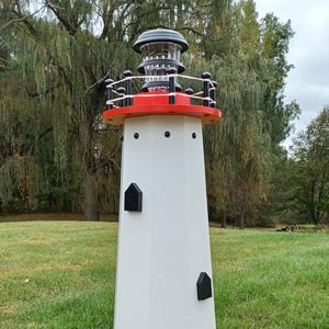 May include: A white, red, and black decorative lighthouse stands on a grassy lawn. The lighthouse features a red top and base, black accents, and a clear, multi-tiered light fixture. The structure has a hexagonal base and is surrounded by green grass.