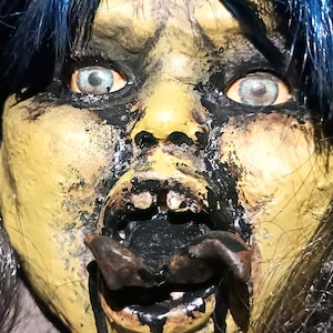 May include: A close-up of a creepy doll's head with blue hair, yellow skin, and black paint around the eyes and mouth. The doll's mouth is open, revealing black paint and a metal hook.