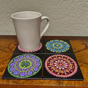 May include: A white ceramic mug rests on a set of four square coasters. Each coaster features a black background with a colorful, hand-painted mandala design. The mandala designs include pink, blue, green, and red patterns.