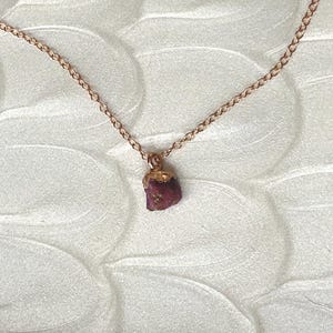 Può includere: Una delicata collana a catena d'oro con un piccolo ciondolo di rubino grezzo. Il rubino è di colore rosso intenso e ha una finitura naturale non lucidata.