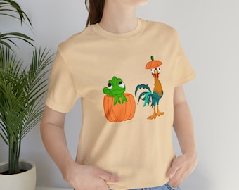 May include: A light beige t-shirt featuring a green chameleon inside a pumpkin and a rooster wearing a pumpkin on its head. The rooster is blue, red, and yellow. The chameleon is green and has a black eye.