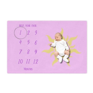 May include: A pink baby milestone blanket with a yellow sun design. The blanket has numbers 1 through 12 printed on it, with the number 1 circled. The text "BEST. YEAR. EVER." is printed at the top of the blanket. A baby is lying on the blanket inside the sun design.