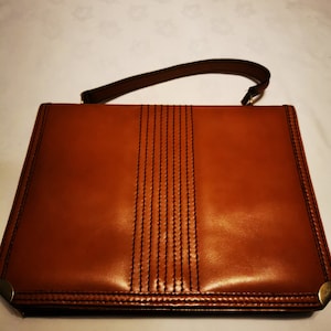 Peut inclure: Un sac à main en cuir marron avec une poignée supérieure et un design cousu. Le sac a une fermeture à rabat et des ferrures dorées.
