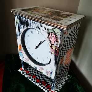 May include: A decorative clock with a white face and black hands. The clock features illustrations of characters from Alice in Wonderland, with a black and white checkered pattern on the sides and red hearts on the base.