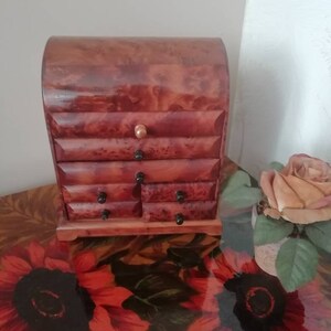 Pode incluir: Uma caixa de joias de madeira com uma tampa curva e seis gavetas. A caixa é feita de madeira escura com um tom avermelhado e tem um padrão de grão de madeira natural. A caixa está sentada em uma mesa com um padrão floral.