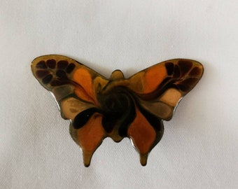 May include: A brown and orange butterfly brooch with black and white accents. The butterfly is made of a translucent material and has a shiny finish.
