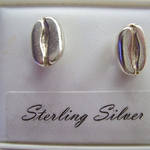 May include: Two sterling silver stud earrings shaped like coffee beans. The earrings are displayed on a white surface inside a white box. The text "Sterling Silver" is printed on a white label.