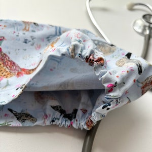 K&ouml;nnte beinhalten: Ein hellblauer Stethoskop-&Uuml;berzug mit wei&szlig;em Hintergrund und einem Muster aus bunten V&ouml;geln und Blumen. Der &Uuml;berzug ist aus Stoff und hat ein Gummiband am Rand.