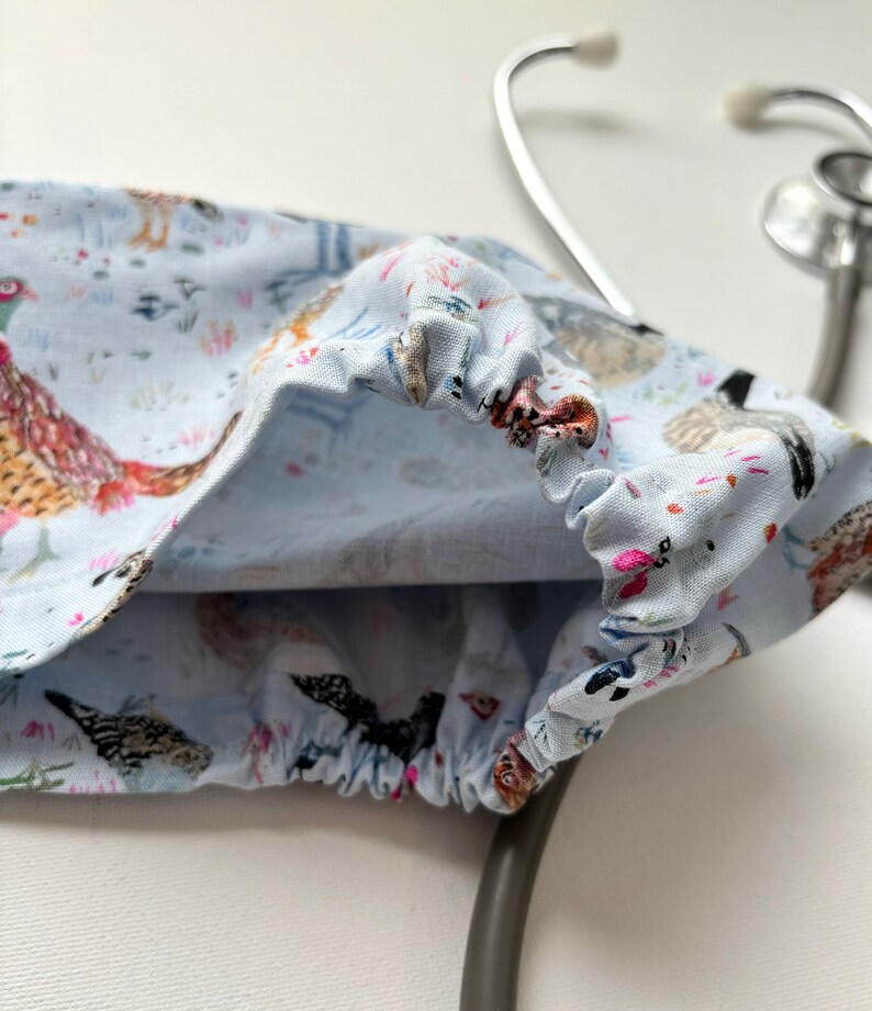 K&ouml;nnte beinhalten: Ein hellblauer Stethoskop-&Uuml;berzug mit wei&szlig;em Hintergrund und einem Muster aus bunten V&ouml;geln und Blumen. Der &Uuml;berzug ist aus Stoff und hat ein Gummiband am Rand.