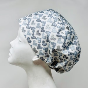 May include: A white scrub cap featuring a repeating pattern of pixelated hearts in shades of grey and blue. The cap is designed to cover the head and hair, commonly worn in medical or care settings.