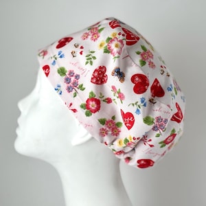 May include: A white scrub hat with a pink floral pattern and red hearts. The hat has the words "I love you" and "so very" printed on it.
