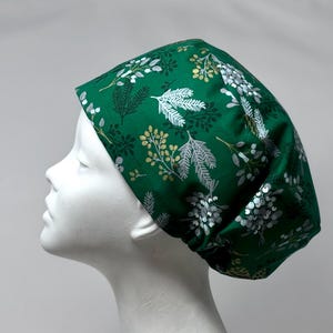 May include: A scrub cap in a deep forest green color featuring a repeating pattern of white and gold botanical illustrations, including pine branches, sprigs, and berries. The cap has a rounded, gathered back design with a fitted front band.