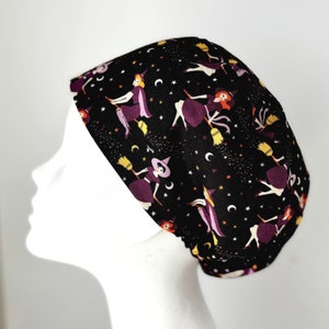 May include: A black scrub hat with a repeating pattern of witches flying on broomsticks against a background of stars and moons. The witches are wearing purple dresses and have orange hair.