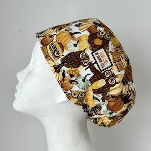 May include: A white scrub hat with a brown, white and orange print of owls and pumpkins. The text "Falling Leaves and Bunnies Await" is printed on the hat.