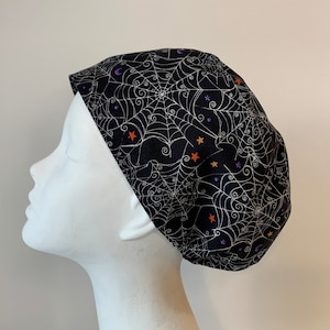 Könnte beinhalten: Schwarze OP-Mütze mit einem weißen Spinnennetz-Muster und orangefarbenen und violetten Sternen.