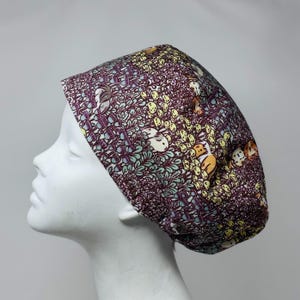 May include: A patterned scrub cap with a repeating design of rabbits, foliage, and other elements in shades of purple, yellow, and green. The cap is displayed on a white mannequin head, showcasing its shape and design.