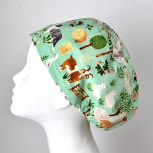 May include: A green scrub hat with a farm animal print. The hat features goats, sheep, a goose, and a hay bale.