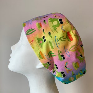 May include: A colourful surgical scrub hat with a pattern of green frogs, lily pads, dragonflies, and reeds on a pink, yellow, and blue ombre background.