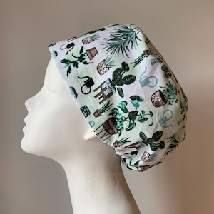 May include: White scrub cap featuring a repeating pattern of potted plants, watering cans, and botanical illustrations in shades of green and brown. The cap is designed to cover the hair and is likely used in a medical or professional setting.