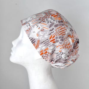 May include: A white scrub hat with a pattern of orange carrots, grey leaves, and white bunnies. The bunnies have black eyes and noses and are smiling.