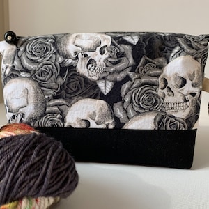 May include: Black and white zippered pouch with a skull and rose print. The pouch has a black fabric handle and a black fabric bottom. The pouch has a small wooden tag that says "A Juggling Raven".