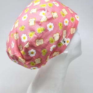 May include: A pink surgical scrub cap featuring a repeating pattern of cartoon eggs, avocado slices, and toast. The cap is made of fabric and is designed to cover the head. The design includes white, yellow, green, and brown elements.