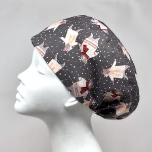 May include: A dark gray scrub cap featuring a repeating pattern of cartoon llamas wearing sweaters and red plaid scarves. The llamas are interspersed with small white polka dots. The cap is shown on a white mannequin head.