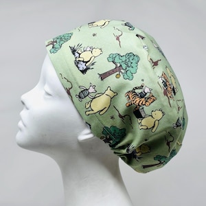 May include: A light green scrub cap featuring a repeating pattern of cartoon characters, including a yellow bear, a tiger, and a pig, with trees and bees. The cap is made of fabric and is designed to cover the head.
