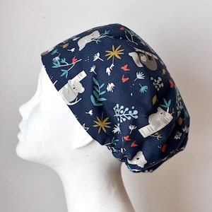 May include: A navy blue scrub cap with a repeating pattern of koalas and sloths. The cap has a white band and is decorated with yellow starbursts, red butterflies, and green foliage. The fabric is a dark blue with a repeating pattern of the animals and plants.