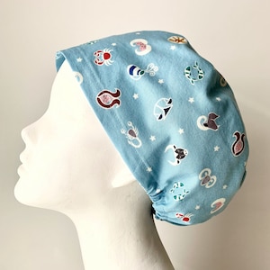 May include: Light blue scrub cap featuring a repeating pattern of zodiac symbols and white stars. The cap is made of fabric and is designed to cover the head. The zodiac signs are in various colors, including red, brown, and white.