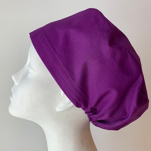 May include: A purple scrub hat made of cotton fabric. The hat has a gathered elastic band at the back.