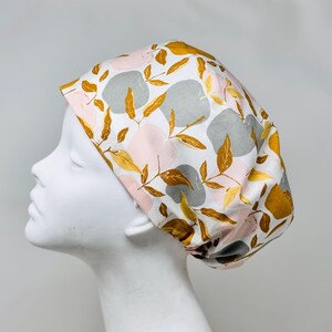 May include: A white scrub cap featuring a pattern of pink, grey, and gold-coloured abstract shapes and leaves. The cap is displayed on a white mannequin head, highlighting the design.