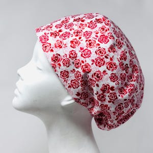May include: A white cap with a repeating pattern of red roses. The roses vary in size and shade, creating a textured look. The cap is made of a soft fabric and is designed to fit snugly on the head.