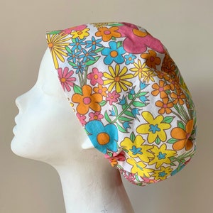 Könnte beinhalten: Eine weiße OP-Mütze mit einem bunten Blumenmuster. Die Blumen sind in den Farben Rosa, Gelb, Blau und Orange gehalten. Die Mütze ist aus einem weichen, bequemen Stoff.