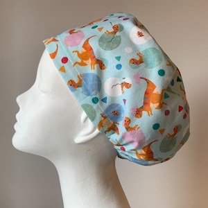 May include: A light blue surgical scrub cap featuring a repeating pattern of orange and brown dinosaurs hatching from eggs. The cap also has colorful geometric shapes. The cap is shown on a white mannequin head.