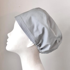 May include: A light gray surgical scrub cap made of fabric. The cap is shown on a white mannequin head. The cap has a band around the forehead and is gathered at the back.