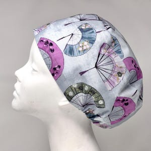 May include: A light gray surgical scrub cap featuring a repeating pattern of Japanese fans in shades of purple, blue, and green. The fans have intricate details and floral accents. The cap is designed to fit snugly on the head.