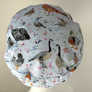 K&ouml;nnte beinhalten: Eine hellblaue Duschhaube aus Stoff mit einem Muster aus verschiedenen V&ouml;geln und Blumen. Zu den V&ouml;geln geh&ouml;ren G&auml;nse, Enten, Wachteln und H&uuml;hner.