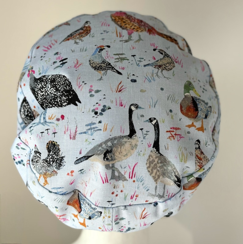 K&ouml;nnte beinhalten: Eine hellblaue Duschhaube aus Stoff mit einem Muster aus verschiedenen V&ouml;geln und Blumen. Zu den V&ouml;geln geh&ouml;ren G&auml;nse, Enten, Wachteln und H&uuml;hner.