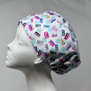 May include: A white scrub cap featuring a repeating pattern of colorful cassette tapes in shades of pink, blue, and grey, interspersed with light blue polka dots. The cap has a rounded, gathered design that fits snugly over the head.