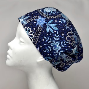 Könnte beinhalten: Eine marineblaue OP-Haube mit einem winterlichen Muster. Die Haube zeigt ein sich wiederholendes Design aus Schneeflocken, Blättern und floralen Elementen in Blau- und Silbertönen. Die Haube ist aus einem weichen Stoff gefertigt.