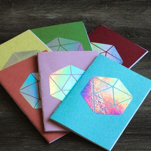 May include: A stack of six colorful notebooks with holographic geometric designs. The notebooks are arranged in a fan shape, with the top notebook being green, followed by yellow, red, purple, blue, and then another blue notebook.
