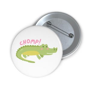 Peut inclure: Un badge blanc avec un dessin de crocodile de dessin animé. Le crocodile est vert avec un ventre jaune et des joues roses. Le mot "CHOMP!" est écrit en rose au-dessus du crocodile. Un deuxième badge avec un dos à épingle argenté est visible.