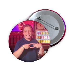 Peut inclure: Un bouton de badge rond avec une personne souriante portant un haut bleu marine. Le badge a un dos argenté et une épingle. L'image sur le badge montre la personne faisant un geste de la main.